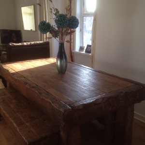 May include: A rustic wooden table with a thick, weathered top and a matching bench. The table is made of dark wood and has a natural, unfinished look. A vase with blue flowers sits on the table.