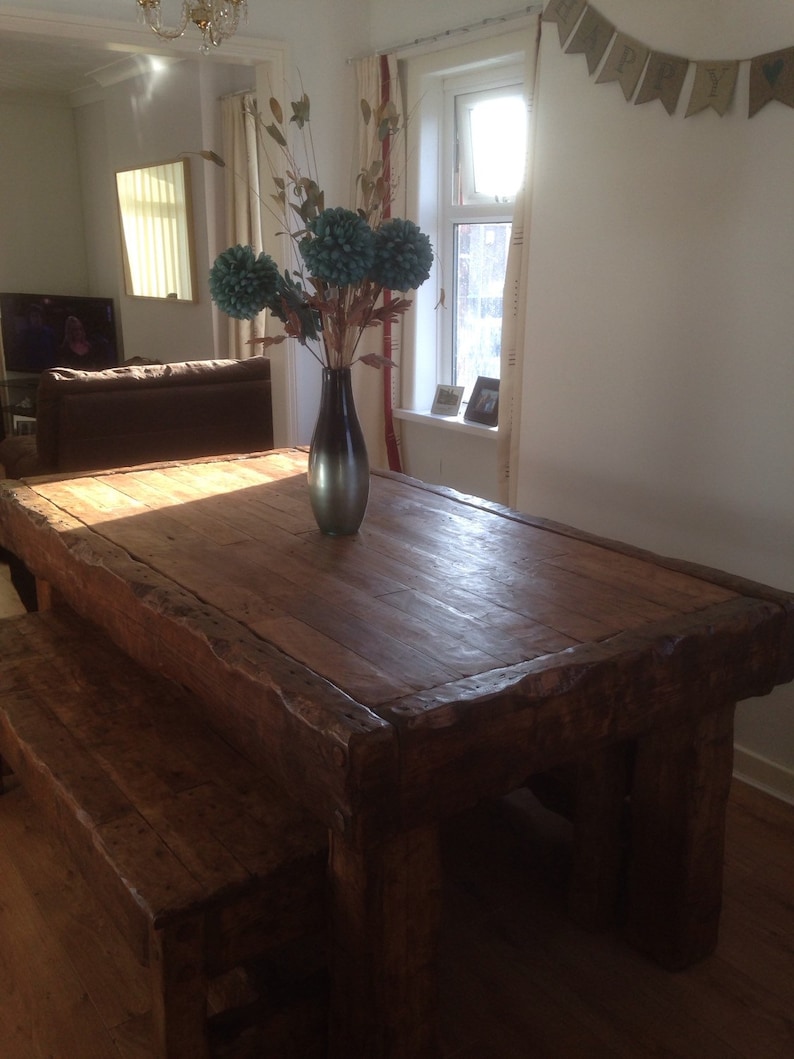May include: A rustic wooden table with a thick, weathered top and a matching bench. The table is made of dark wood and has a natural, unfinished look. A vase with blue flowers sits on the table.