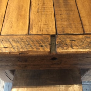 May include: Close-up of a rustic wooden table. The tabletop is constructed from several planks of light brown wood, with visible grain and knots. The edges of the table have a darker, weathered appearance. The table's base is also made of wood, providing a sturdy foundation.
