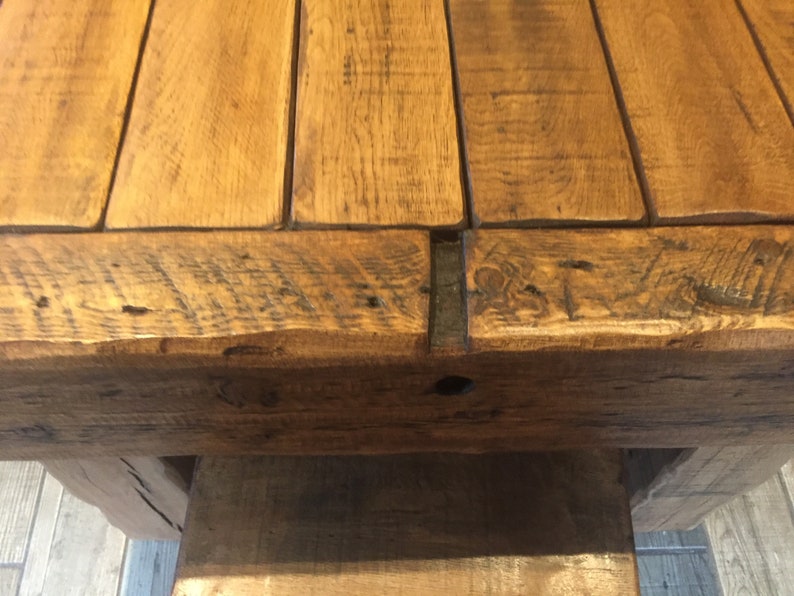 May include: Close-up of a rustic wooden table. The tabletop is constructed from several planks of light brown wood, with visible grain and knots. The edges of the table have a darker, weathered appearance. The table's base is also made of wood, providing a sturdy foundation.