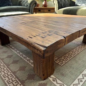 May include: A low, square coffee table constructed from thick, dark brown wooden planks. The surface features visible wood grain, natural knots, and four prominent circular wooden dowels at each corner, supported by four sturdy, block-style legs.