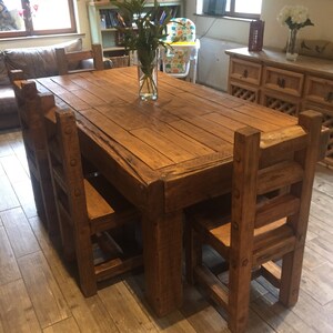 May include: A rustic wooden dining table with six matching chairs. The table is rectangular and has a natural wood finish. A clear glass vase with green foliage sits in the center. The chairs have a ladder-back design.