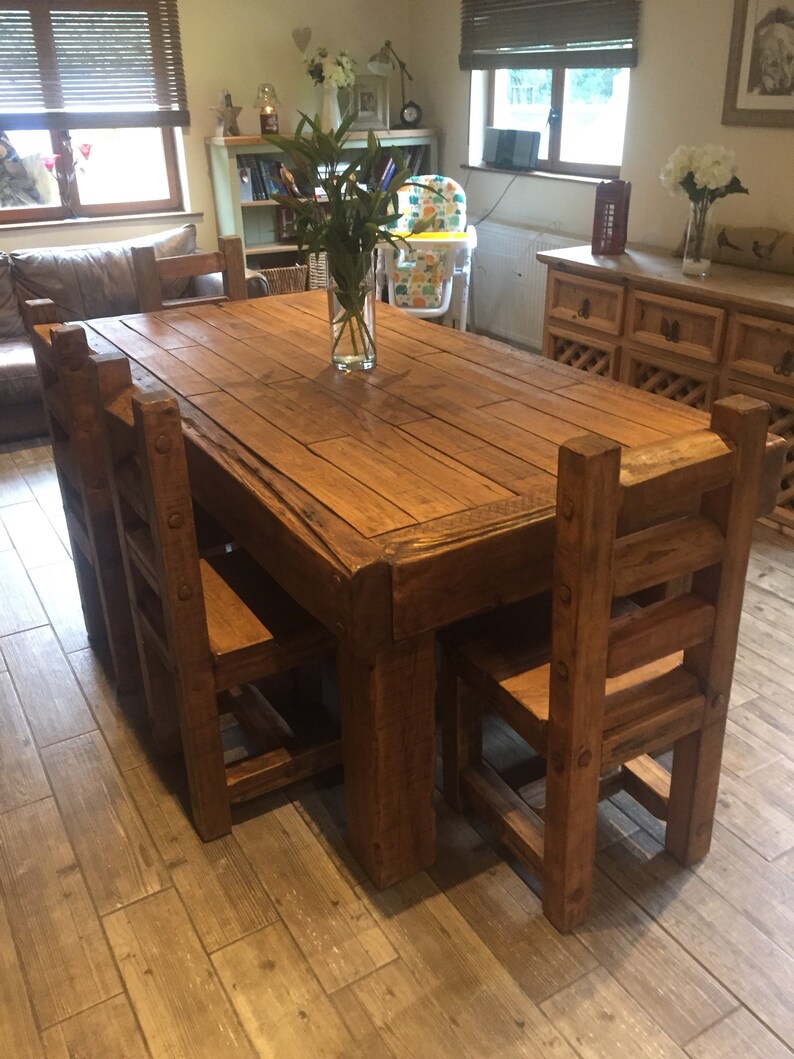May include: A rustic wooden dining table with six matching chairs. The table is rectangular and has a natural wood finish. A clear glass vase with green foliage sits in the center. The chairs have a ladder-back design.