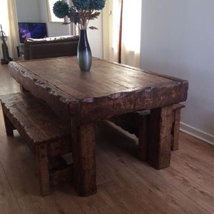 May include: A rustic wooden dining table with a thick, uneven top and two matching benches. The table is made of reclaimed wood and has a natural, distressed finish. The table is set in a room with hardwood floors and a vase of blue flowers on the table.