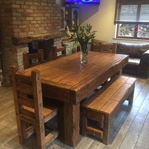 May include: A rustic wooden dining table with matching chairs and a bench. The table is rectangular and has a natural wood finish. The chairs have a ladder-back design. The bench is placed on one side of the table. A vase of green plants sits on the table.