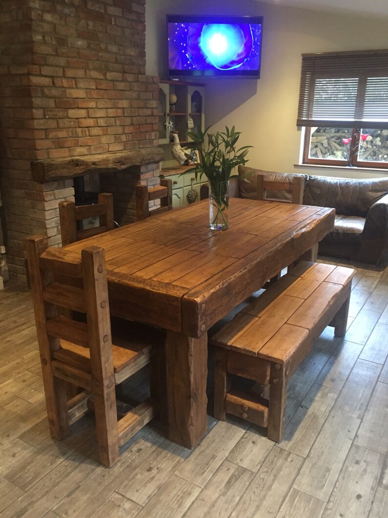 May include: A rustic wooden dining table with matching chairs and a bench. The table is rectangular and has a natural wood finish. The chairs have a ladder-back design. The bench is placed on one side of the table. A vase of green plants sits on the table.