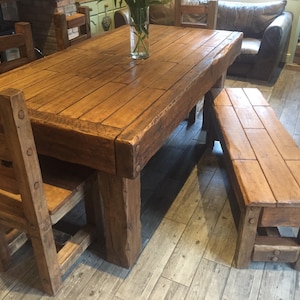 May include: A rustic wooden dining set featuring a large rectangular table, a bench, and two chairs. The table and bench are made of thick, solid wood with a natural finish. The chairs have a ladder-back design. The set is ideal for a dining room.