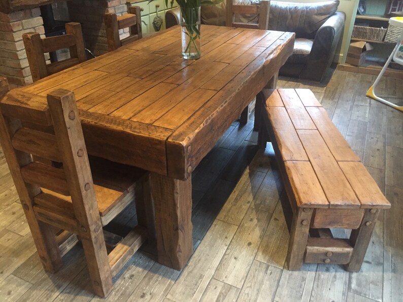 May include: A rustic wooden dining set featuring a large rectangular table, a bench, and two chairs. The table and bench are made of thick, solid wood with a natural finish. The chairs have a ladder-back design. The set is ideal for a dining room.