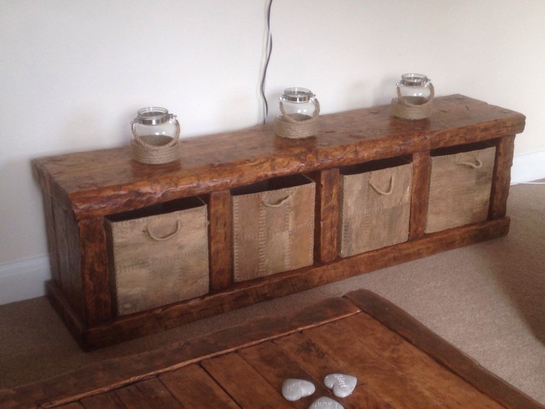 May include: A rustic wooden bench with four storage baskets. The bench is stained a dark brown color and has a natural wood finish. The baskets are made of woven material and have a natural color. There are three glass candle holders on top of the bench.