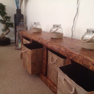 May include: A rustic wooden console table with four woven baskets and three glass jars with rope accents. The table has a natural wood finish and is decorated with a small bonsai tree and a Buddha statue.