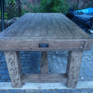 May include: A rustic wooden table with a weathered finish. The table has a thick, rectangular top and sturdy legs. A small metal plaque on the table reads "Beautifully Handcrafted In The UK By Rita Long Island Furniture".