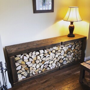 May include: A rustic wooden log storage rack with a dark brown finish. The rack is filled with stacked pieces of firewood, creating a cozy and inviting atmosphere. A lamp with a white shade sits on top of the rack.