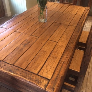May include: A large, rectangular wooden dining table with a natural finish. The tabletop is constructed from planks of wood, and a clear glass vase with green stems sits in the center. The table is surrounded by wooden chairs.