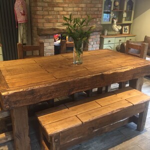 May include: A large, rustic wooden dining table with a matching bench and chairs. The table is made of thick, reclaimed wood and has a natural, unfinished look. A clear glass vase with green stems sits in the center of the table. The table measures approximately 96 inches long.
