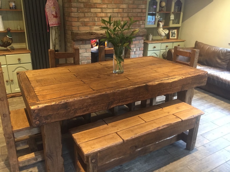 May include: A large, rustic wooden dining table with a matching bench and chairs. The table is made of thick, reclaimed wood and has a natural, unfinished look. A clear glass vase with green stems sits in the center of the table. The table measures approximately 96 inches long.