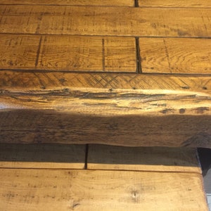 May include: Close-up of a rustic wooden table. The tabletop is constructed from wide, light brown planks with visible wood grain and dark accents. The table's edge and support structure are also visible, showcasing the natural wood texture.