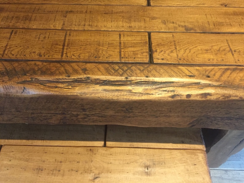May include: Close-up of a rustic wooden table. The tabletop is constructed from wide, light brown planks with visible wood grain and dark accents. The table's edge and support structure are also visible, showcasing the natural wood texture.