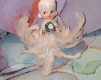 May include: A vintage Christmas tree topper featuring a porcelain doll with a white feathered skirt and holding a small wreath. The doll has blonde hair and rosy cheeks. The wreath is green with red berries.