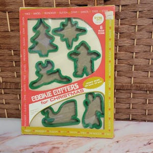 May include: A vintage box of green plastic cookie cutters for Christmas. The box has a red background with white lettering and a picture of the six cookie cutters: a tree, a star, a reindeer, a snowman, a sleigh, and an angel. The box says "Cookie Cutters for Christmas" and "6 Holiday Designs".