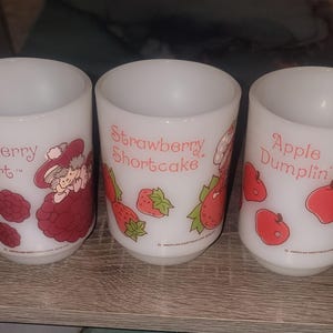 May include: Three white milk glass tumblers featuring Strawberry Shortcake characters. The first cup shows Raspberry Tart with raspberries, the second shows Strawberry Shortcake with strawberries, and the third shows Apple Dumplin with red apples.