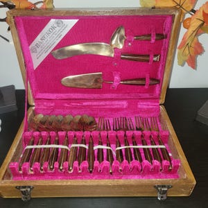May include: A vintage wooden cutlery set in a pink velvet-lined case. The set includes spoons, forks, knives, and serving utensils with gold-colored blades and dark wood handles. The case has a metal clasp and a label that reads "Bangkok".