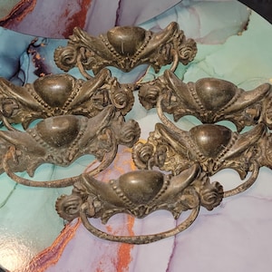 May include: Six antique bronze drawer pulls with ornate, floral designs. Each pull features a central heart-shaped element and a curved handle. The pulls are arranged on a marbled surface, showcasing their aged patina and intricate details.
