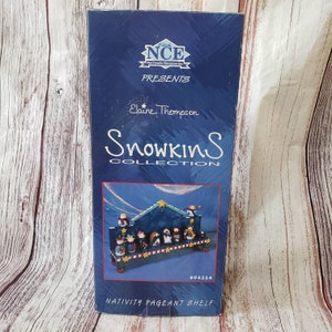 May include: A blue and white wooden nativity scene shelf with a blue background and gold stars. The shelf is labeled "Snowkins Collection" and "Nativity Pageant Shelf".