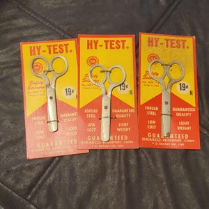 May include: Three vintage HY-TEST safety scissors on their original yellow and red cardboard packaging. The packaging features the text "HY-TEST," "Safety Scissors," and "Forged Steel." Each pair of scissors is priced at 19¢.