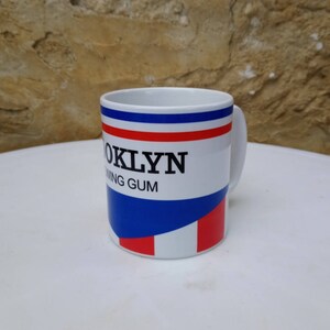 May include: White ceramic mug with a blue, red, and white striped design. The mug has the text "Brooklyn Chewing Gum" printed on it.