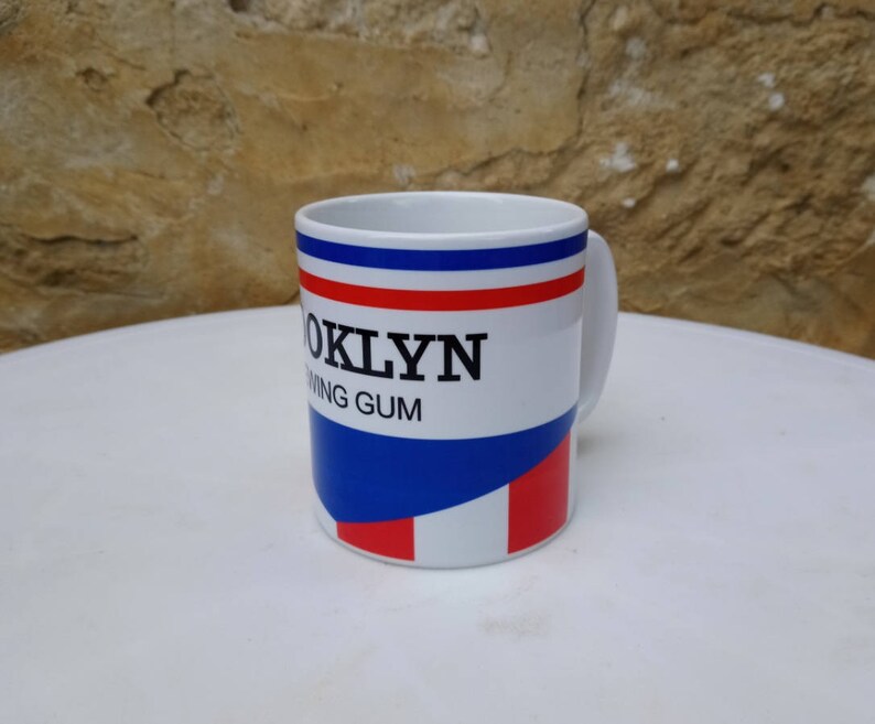 May include: White ceramic mug with a blue, red, and white striped design. The mug has the text "Brooklyn Chewing Gum" printed on it.