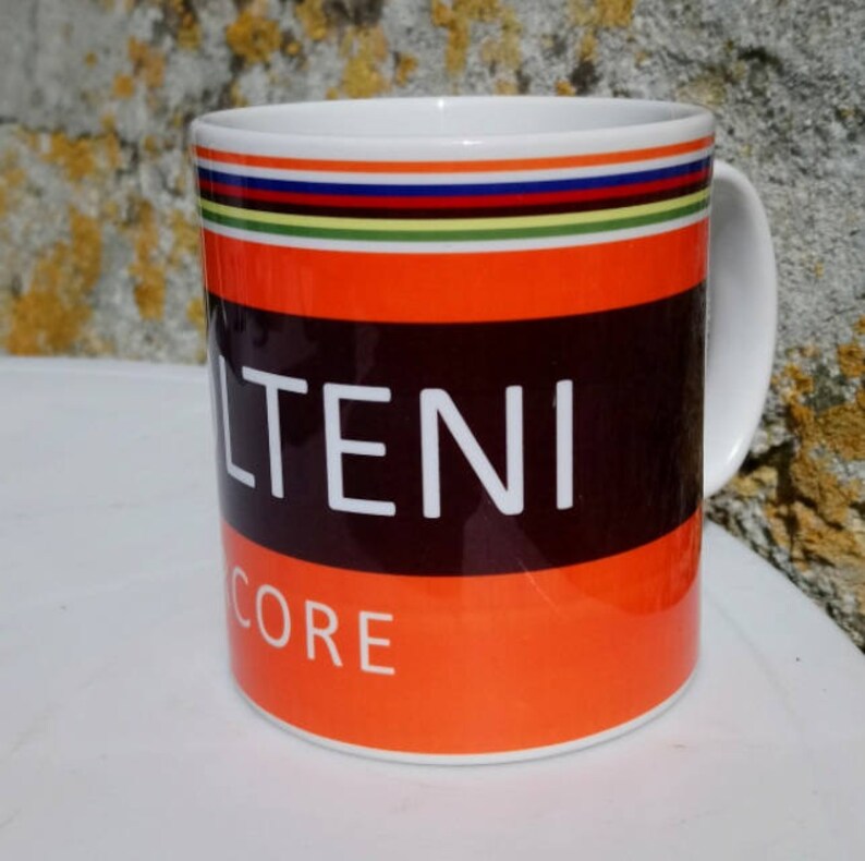 May include: White ceramic mug with a black and orange design. The mug has a black band with the word "ALTENI" in white letters. Below the band is an orange band with the word "CORE" in white letters. The top of the mug has a multi-colored stripe.