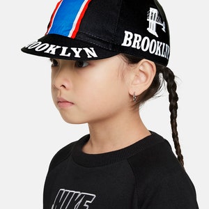 May include: Black cycling cap with a red, white, and blue stripe down the side. The cap has "BROOKLYN" embroidered on the front and a white graphic of the Brooklyn Bridge on the side.