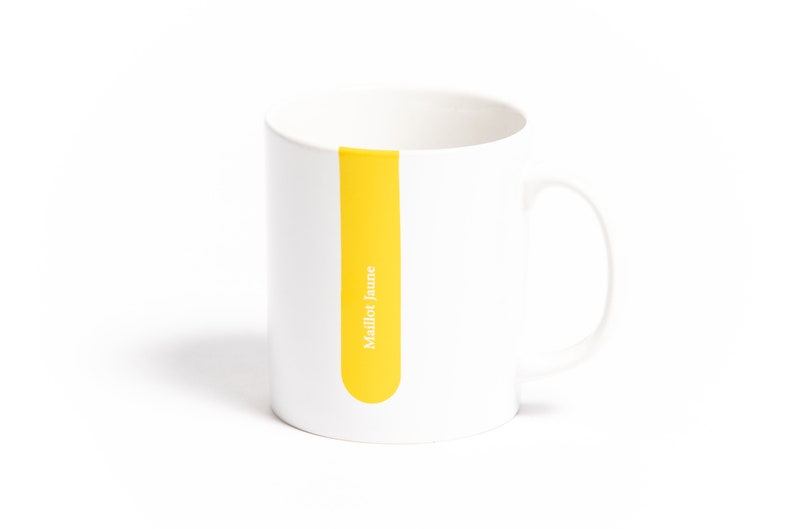 Op de afbeelding: Witte keramische mok met een gele streep en de tekst "Maillot Jaune" erop gedrukt.