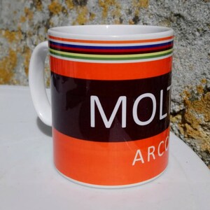 May include: White ceramic mug with a black and orange design. The mug has a black band with the word "MOLTEN" in white letters. Below the band is an orange band with the word "ARCO" in white letters. The top of the mug has a multi-colored stripe design.