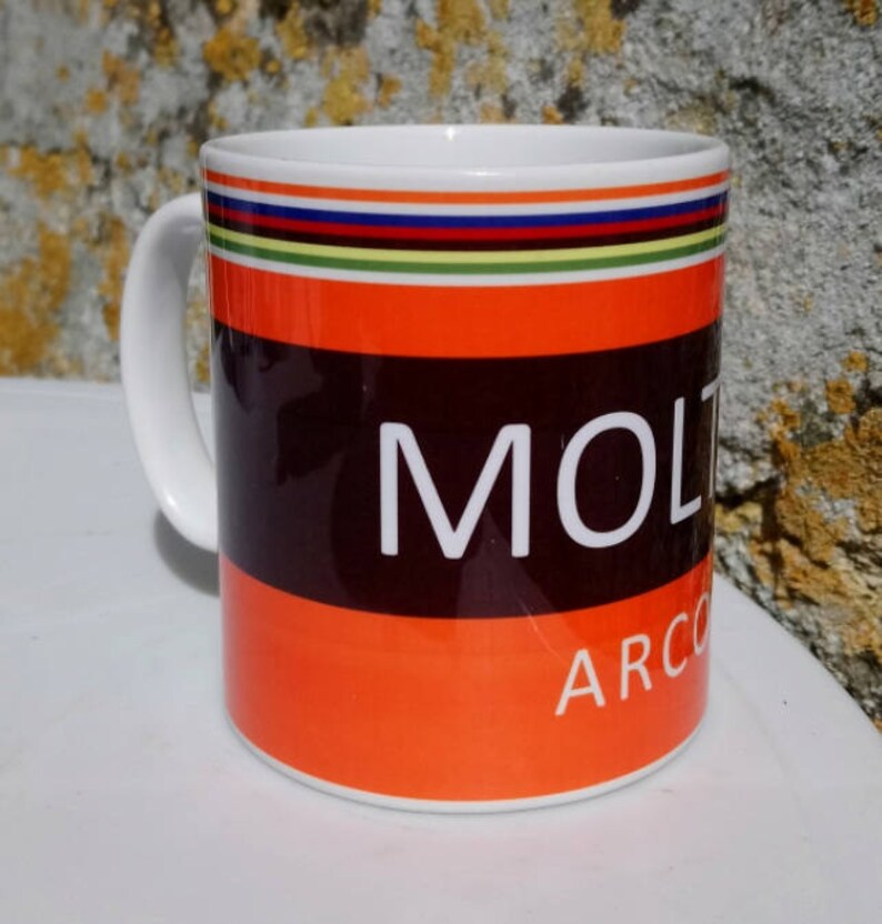 May include: White ceramic mug with a black and orange design. The mug has a black band with the word "MOLTEN" in white letters. Below the band is an orange band with the word "ARCO" in white letters. The top of the mug has a multi-colored stripe design.