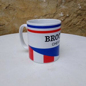May include: White ceramic mug with a blue, red, and white striped design. The mug has the text "BROCK CHEWING" printed on it.