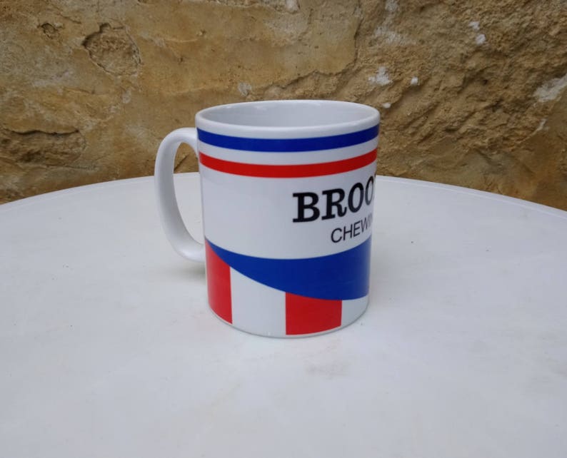 May include: White ceramic mug with a blue, red, and white striped design. The mug has the text "BROCK CHEWING" printed on it.