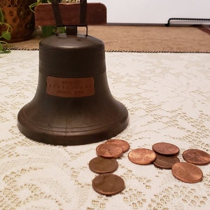 May include: A vintage brass bell with the inscription "World's Standard" and "Made in USA" on a white lace tablecloth with copper-colored coins scattered around it.