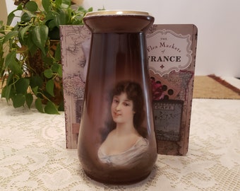 May include: A brown ceramic vase with a painted portrait of a woman with brown hair. The vase has a gold rim and is decorated with a floral design.