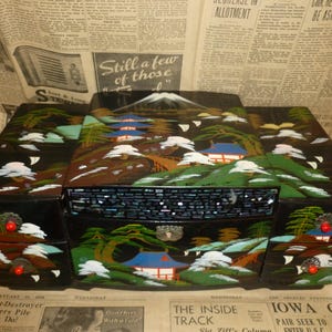 May include: A black lacquered jewelry box with a Japanese landscape design. The box features a mountain, trees, and buildings with blue roofs. The box has a drawer and hinged compartments. Red accents and white clouds are also visible.