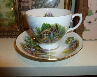 May include: A white teacup and saucer with a gold rim. The teacup and saucer feature a cottage scene with a thatched roof and a garden.