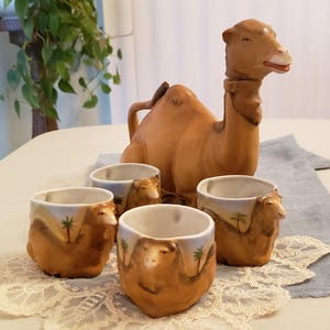 May include: A vintage ceramic camel-shaped decanter with four matching cups. The cups and decanter are tan with painted desert scenes. The set is displayed on a white lace doily and a blue runner.