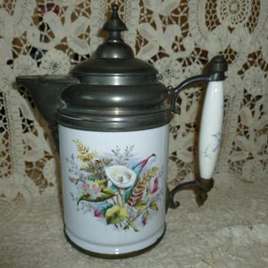 May include: Vintage white ceramic beer stein with a pewter lid and handle. The stein features a floral design with white calla lilies, green leaves, and colorful accents. The handle is white with a floral design.