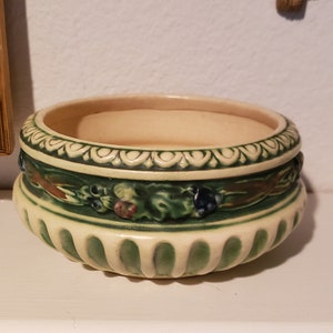 May include: Cream-colored ceramic bowl with a green and floral design. The bowl has a wide, shallow shape with a decorative band of green and floral accents around the middle. The rim has a decorative pattern.