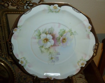 May include: A white porcelain serving platter with a gold rim and floral design. The platter features a central motif of white roses with pink and green accents. The rim is decorated with a repeating pattern of gold accents.
