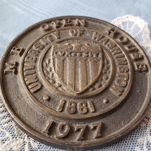 May include: A round, bronze-colored metal medallion with raised lettering and a central emblem. The text reads "M.B. UNIVERSITY OF WASHINGTON" and includes the years "1861" and "1977". The emblem features a shield design.