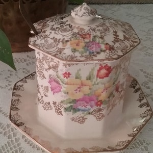 May include: White ceramic sugar bowl with a lid and a matching saucer. The bowl and saucer are decorated with a floral pattern in shades of pink, yellow, and green. The floral pattern is surrounded by a gold trim.