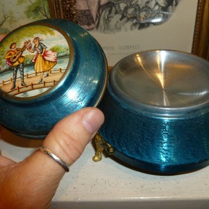 May include: A blue metal music box with a lid featuring a painted scene of two people on a bridge. The lid is closed and the music box is sitting on a white surface.