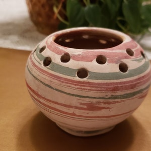 May include: A small, round, white ceramic vase with pink and green stripes and holes around the top. The vase is sitting on a brown surface.