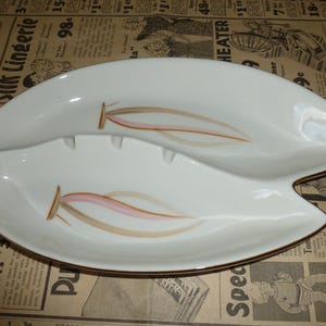 May include: A white ceramic dish shaped like a stylized leaf with two distinct sections. The surface features two identical graphic designs in soft pink and tan colors, each depicting a slender, elongated form. The dish has a smooth, glossy finish.